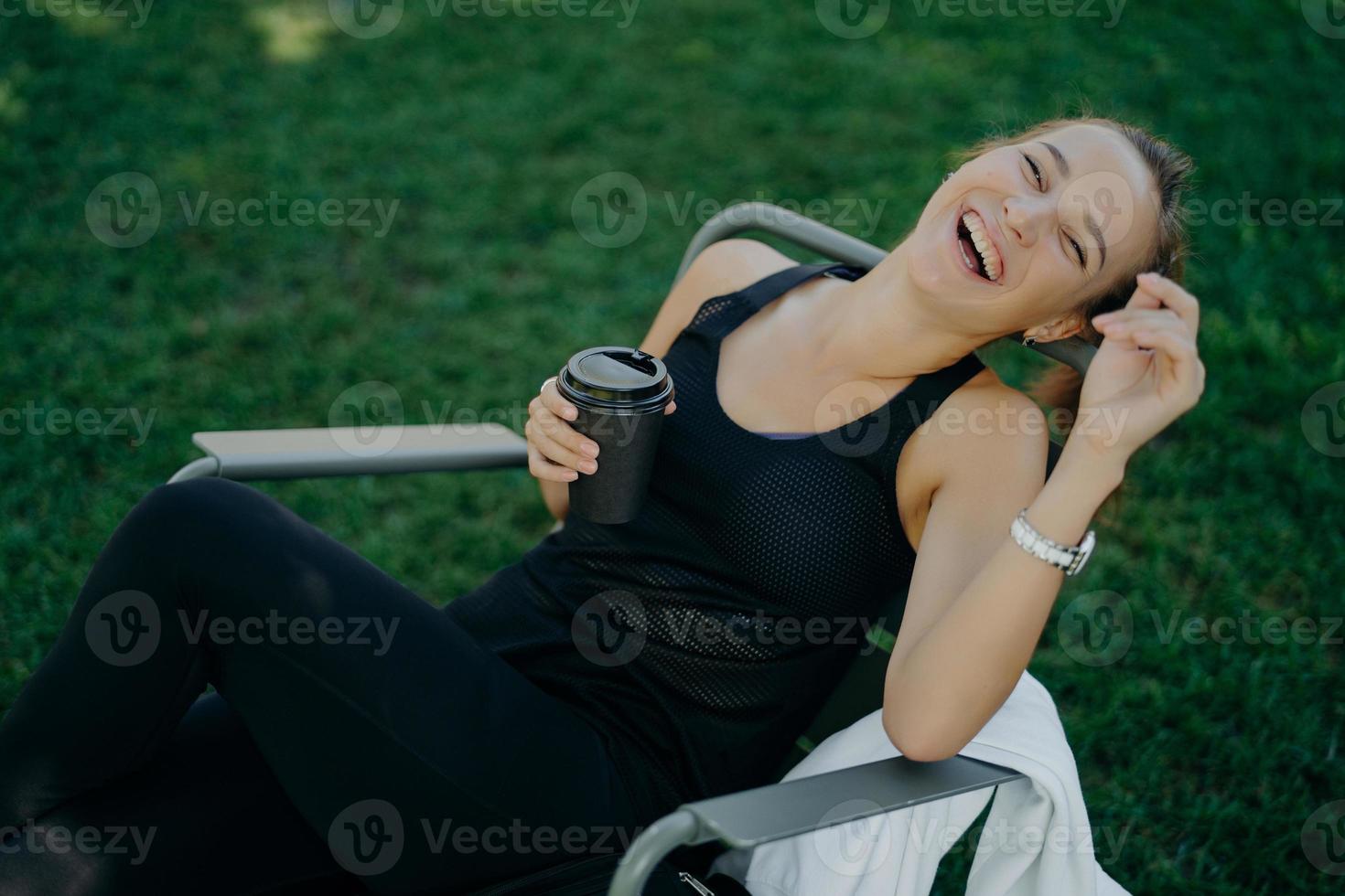 Overjoyed relaxed woman smiles broadly enjoys drinking coffee outdoor dressed in t shirt and trousers poses in comfortable chair against green grass. View from above. People leisure lifestyle photo