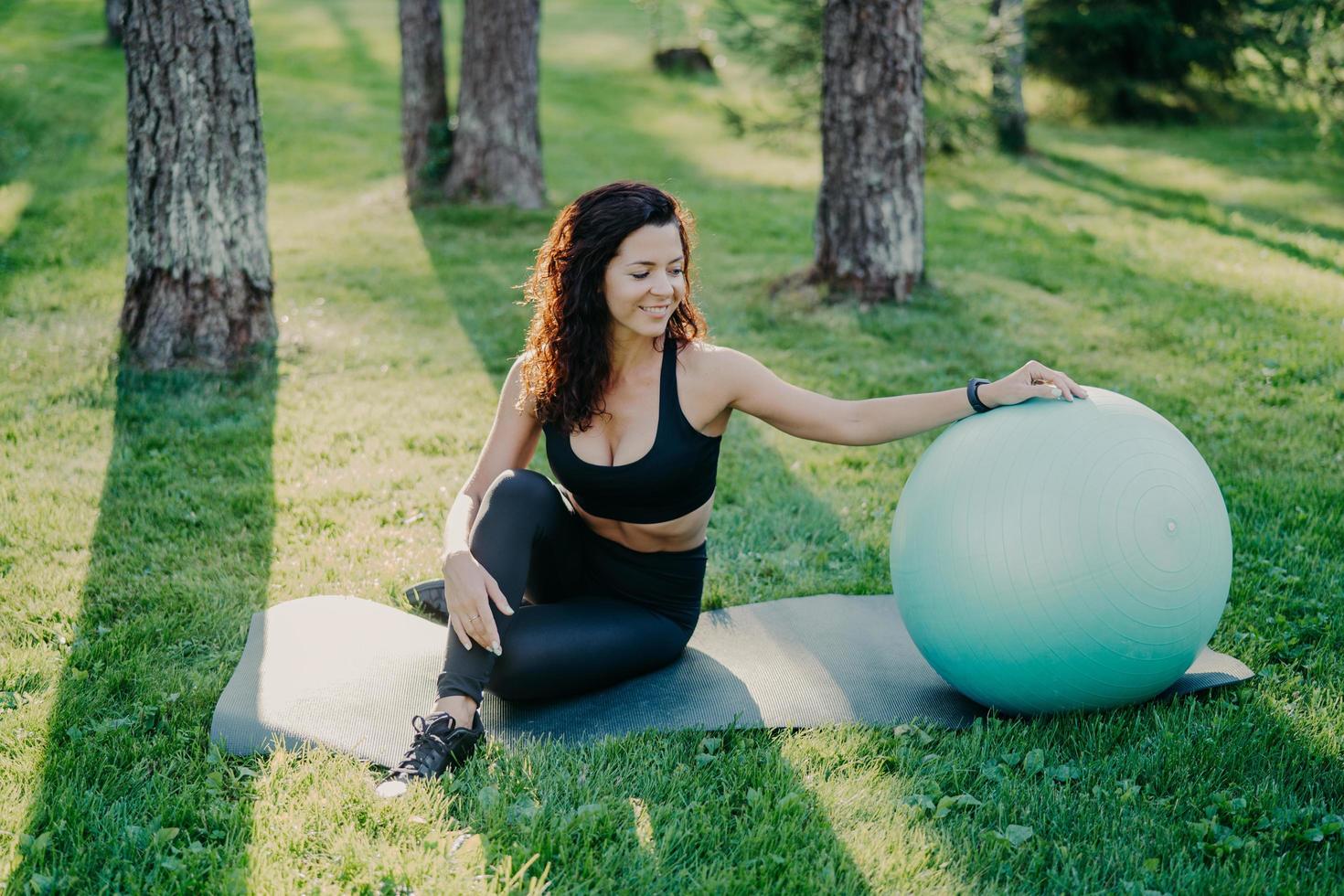 Outdoor shot of active sporty woman takes break after physical exercises, poses on fitness mat with fitball, enjoys fresh air in forest, has perfect body shape uses sport equipment. Gymnastics concept photo