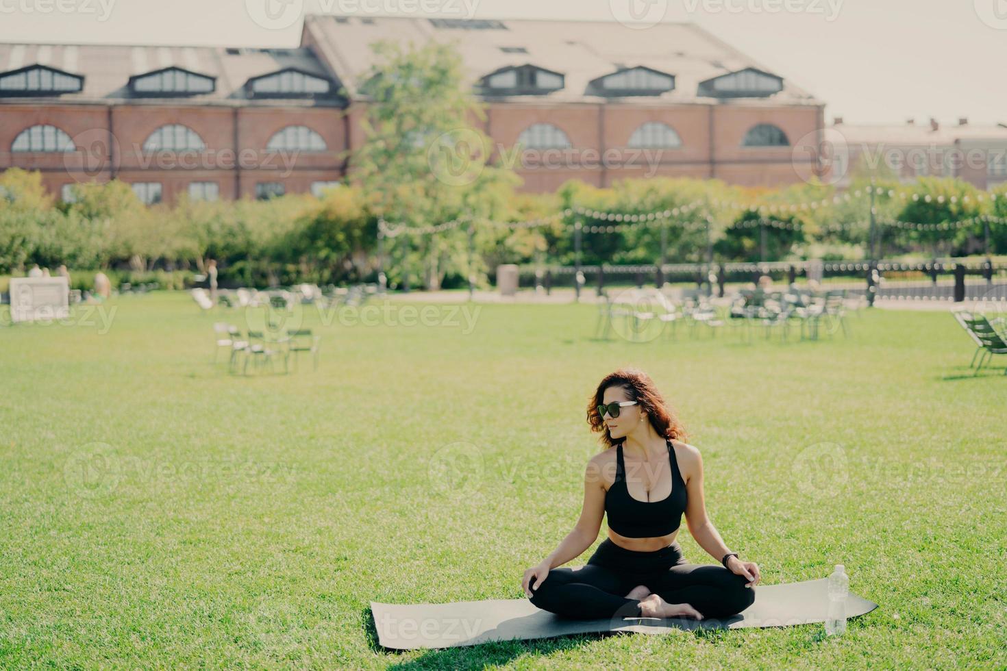 Relaxed slim brunette Caucasian woman sits in lotus pose on fitness mat has perfect body shape does yoga and meditation wears sports clothes poses outdoor. Health fitness and relaxation concept photo