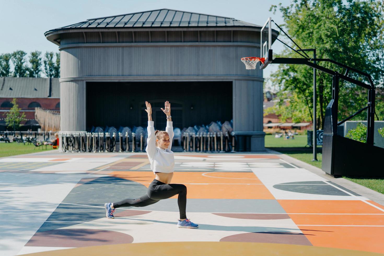 Personal workout routine. Athletic woman does physical exercises outdoor keeps arms raised up warms up before jogging dressed in active wear poses on basketball court. Flexibility and fitness photo