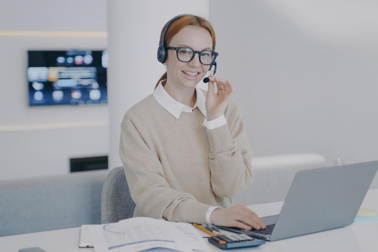 Online interpreter. Young happy woman in headset is talking to client in front of laptop. photo