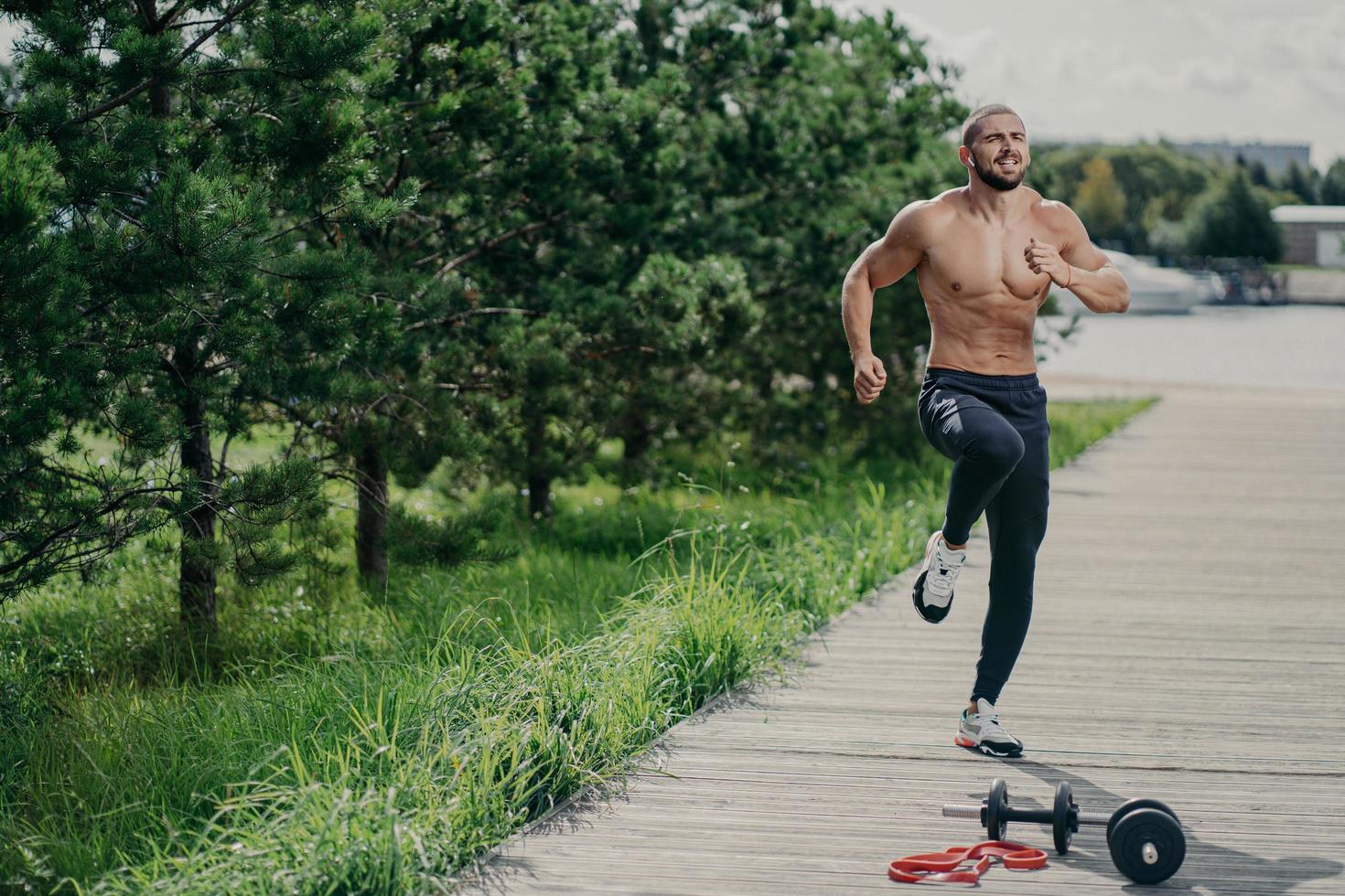 Photo of healthy motivated man with thick bristle, exercises outdoor, jumps and leads healthy lifestyle, poses near sport equipment, has strong muscular body. Fitness, sport and exercising concept