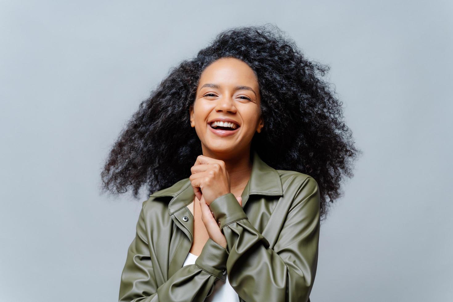 Overjoyed dark skinned curly woman laughs happily, laughs at funny joke, keeps hands pressed together, dressed in fashionable clothes, isolated over grey background. People and positiveness. photo