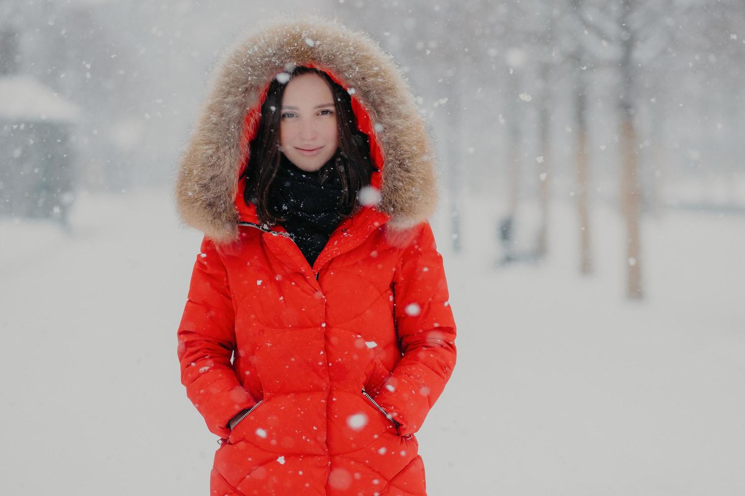 Good looking young woman has appealing look, wears warm red jacket, keeps hands in pockets, has outdoor stroll during winter time and snowfall, poses over blurred snow background. Season concept photo