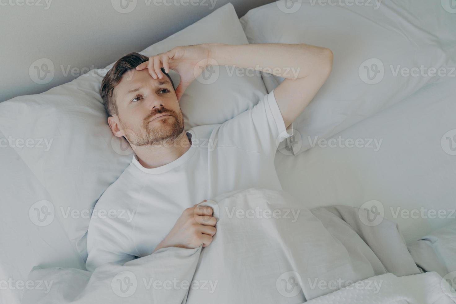Young tired bearded man lying on bed with open eyes photo
