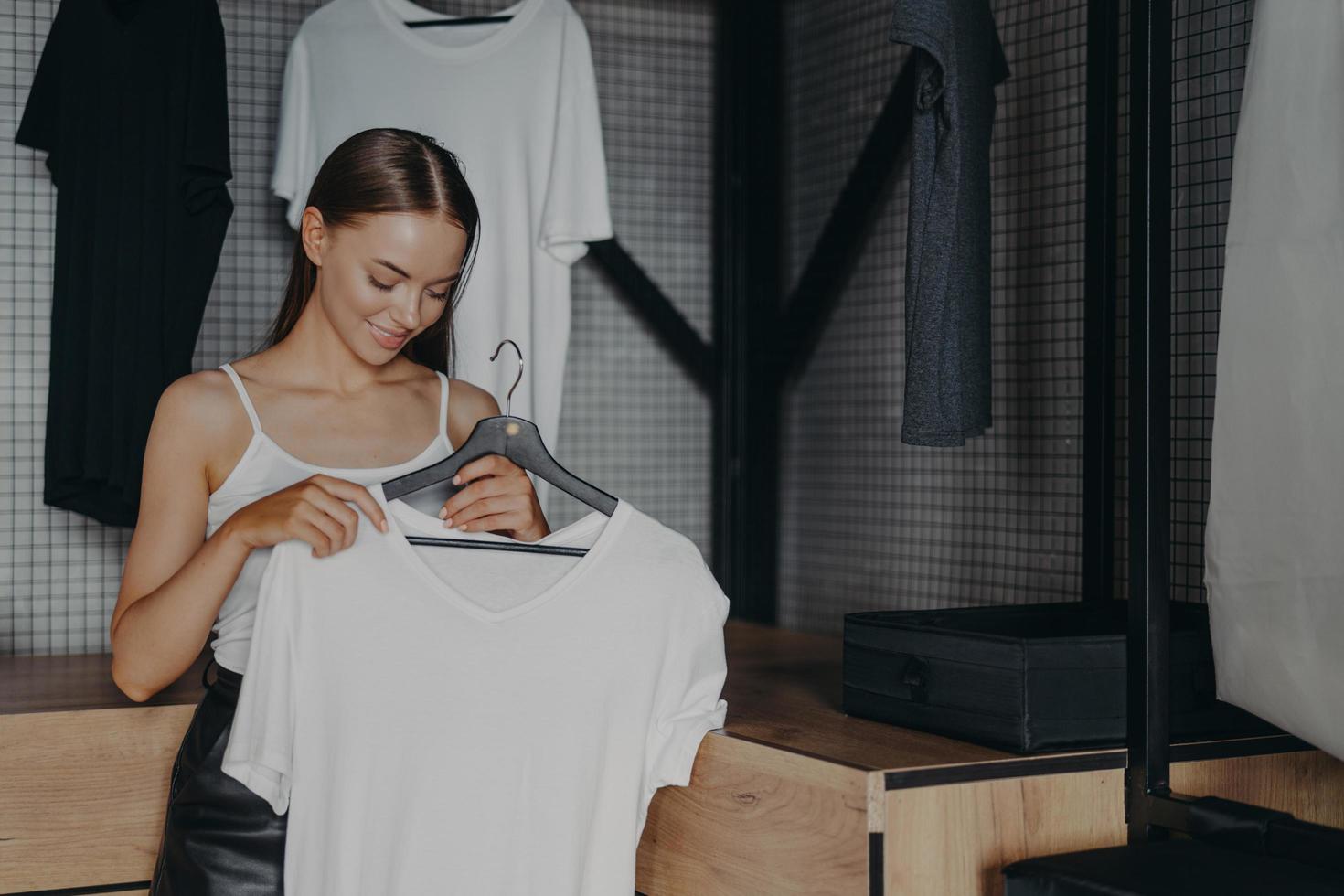Brunette young woman chooses outfit from home wardrobe, holds white blouse on hangers, picks outfit to wear on specical occasion, poses indoor. Women, dressing, clothing and fashion concept. photo