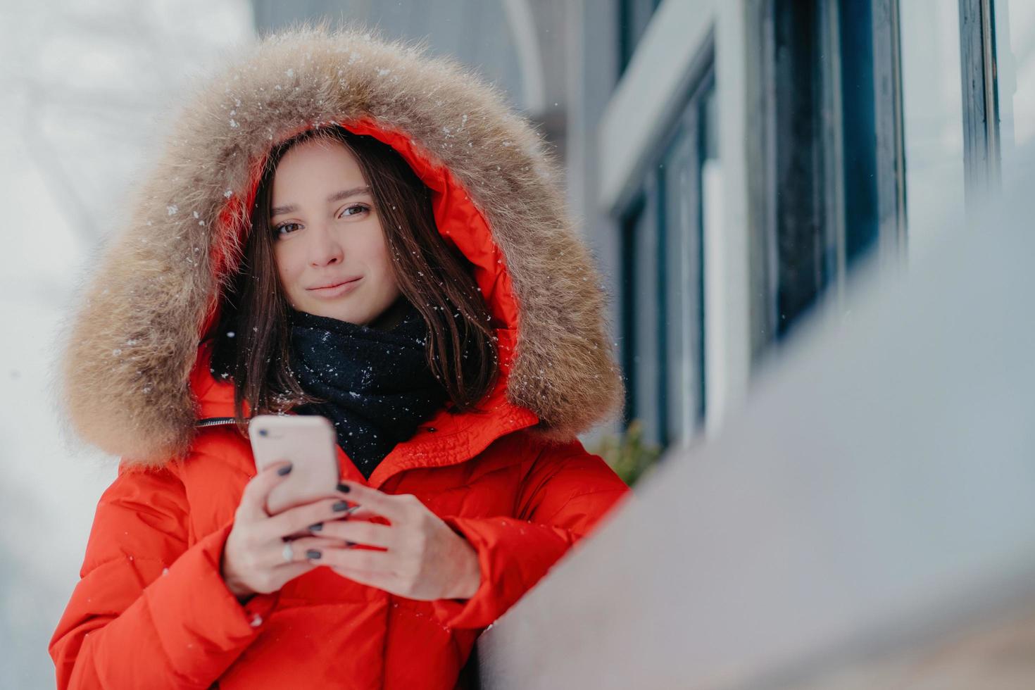 Photo of attractive woman holds modern modern gadget, wears red jacket with hoody, checks email box, connected to wireless internet, stands outdoor, enjoys free time and frosty winter weather