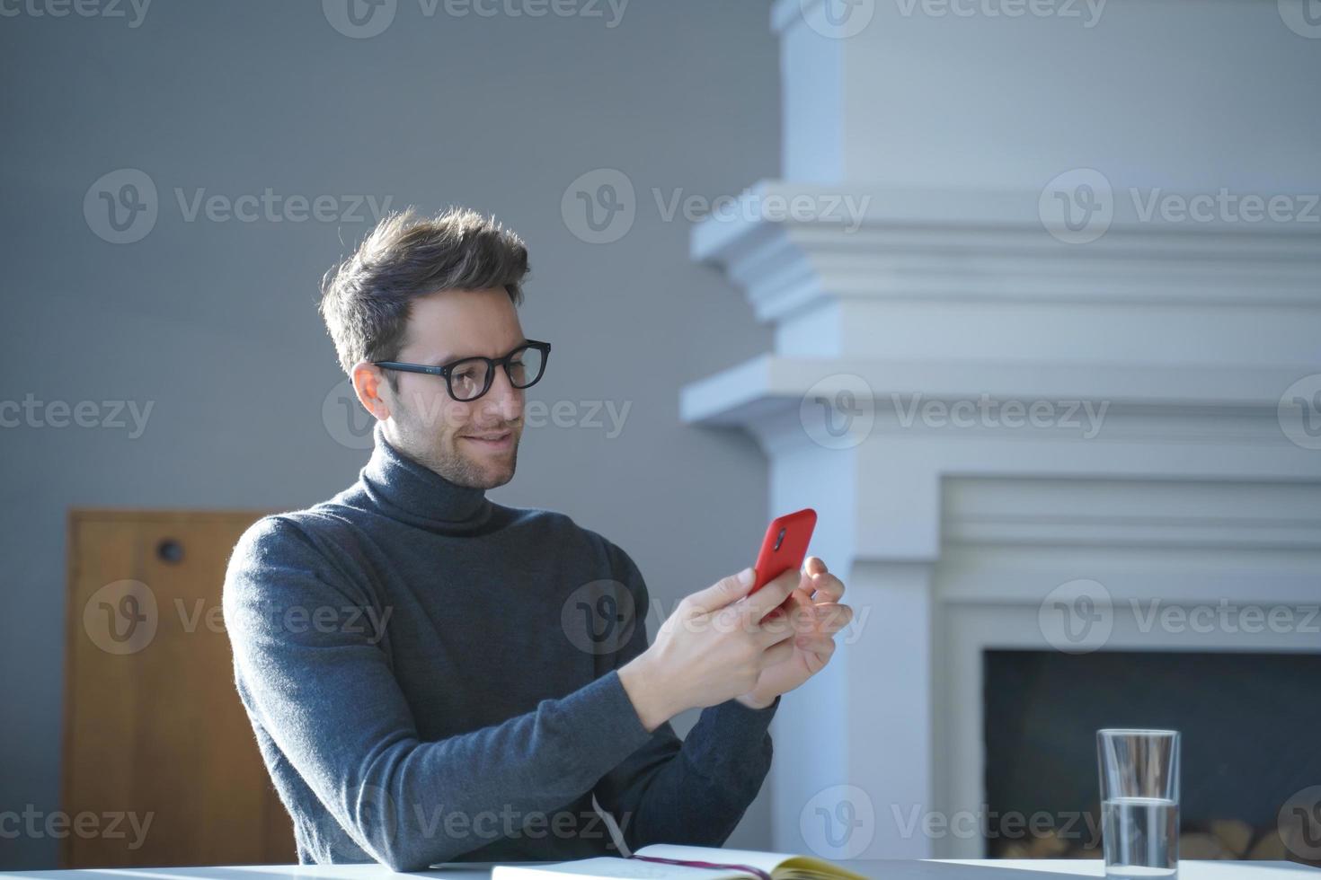 Young handsome german man in glasses sitting at table and using mobile phone at home photo