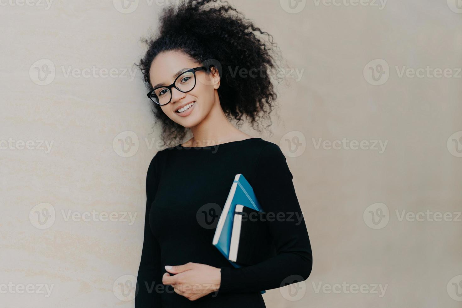 Positive curly African American student carries notepad and diary, wears black clothes, transparent glasses, enjoys studying in university, isolated on grey background. Cute woman with notebook photo