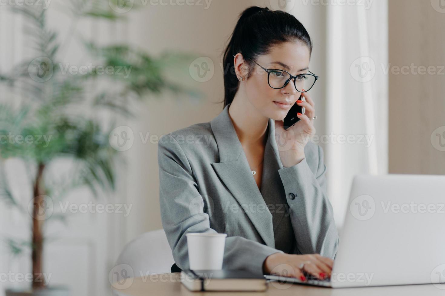 Serious brunette woman keyboards information, concentrated into laptop computer, dressed in formal outfit, poses at coworking space, works remotely, involved in working process during daytime. photo