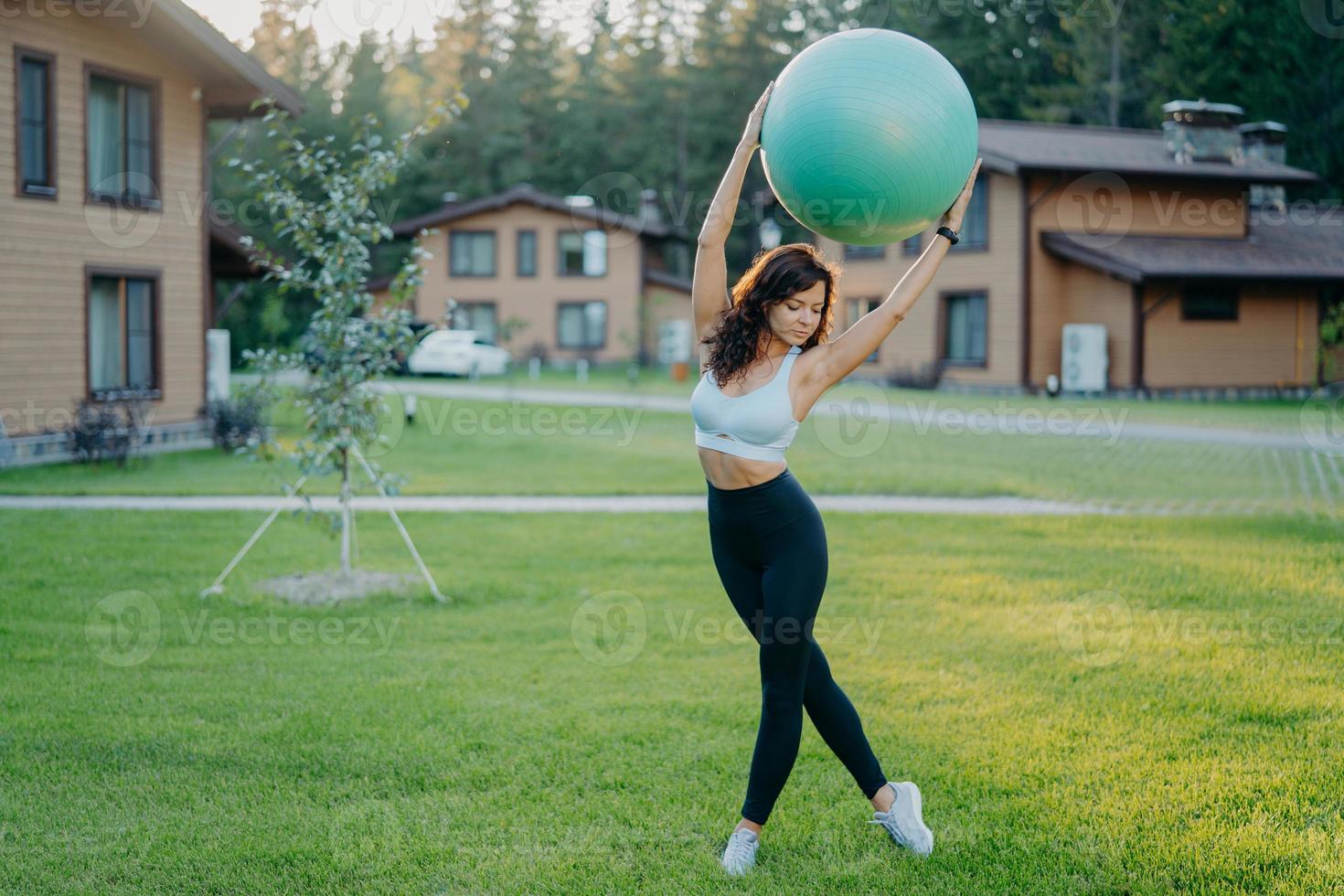 Slim young European woman holds fitball over head, dressed in cropped top and leggings, has gymnastics exercises outdoor, poses at green lawn near private house. People, sport, training concept photo