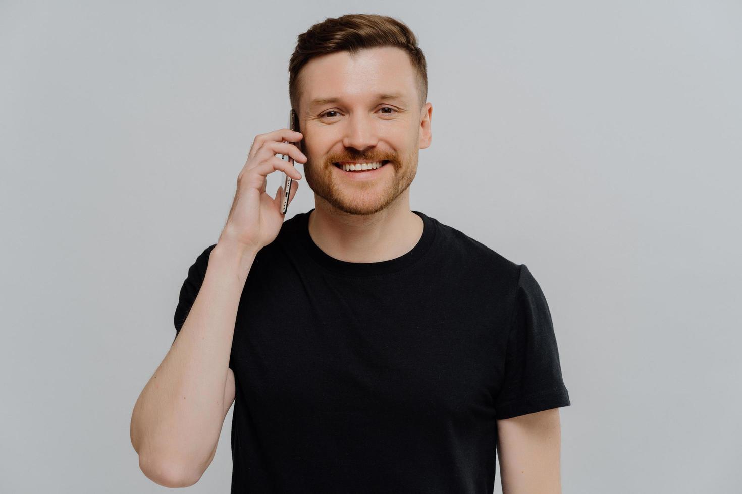 Handsome adult man makes call holds smartphone near ear enjoys pleasant conversation with friend dressed in casual black t shirt isolated over blue background glad to hear girlfriends voice. photo