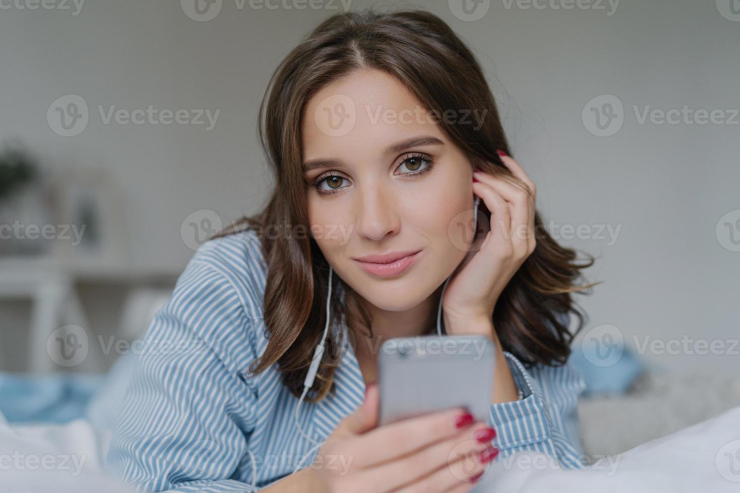 Close up shot of attractive brunette young woman downloads songs from network, uses smart phone for entertainment, dressed in nightclothes, rests in bed, has lazy day, looks seriously at camera photo