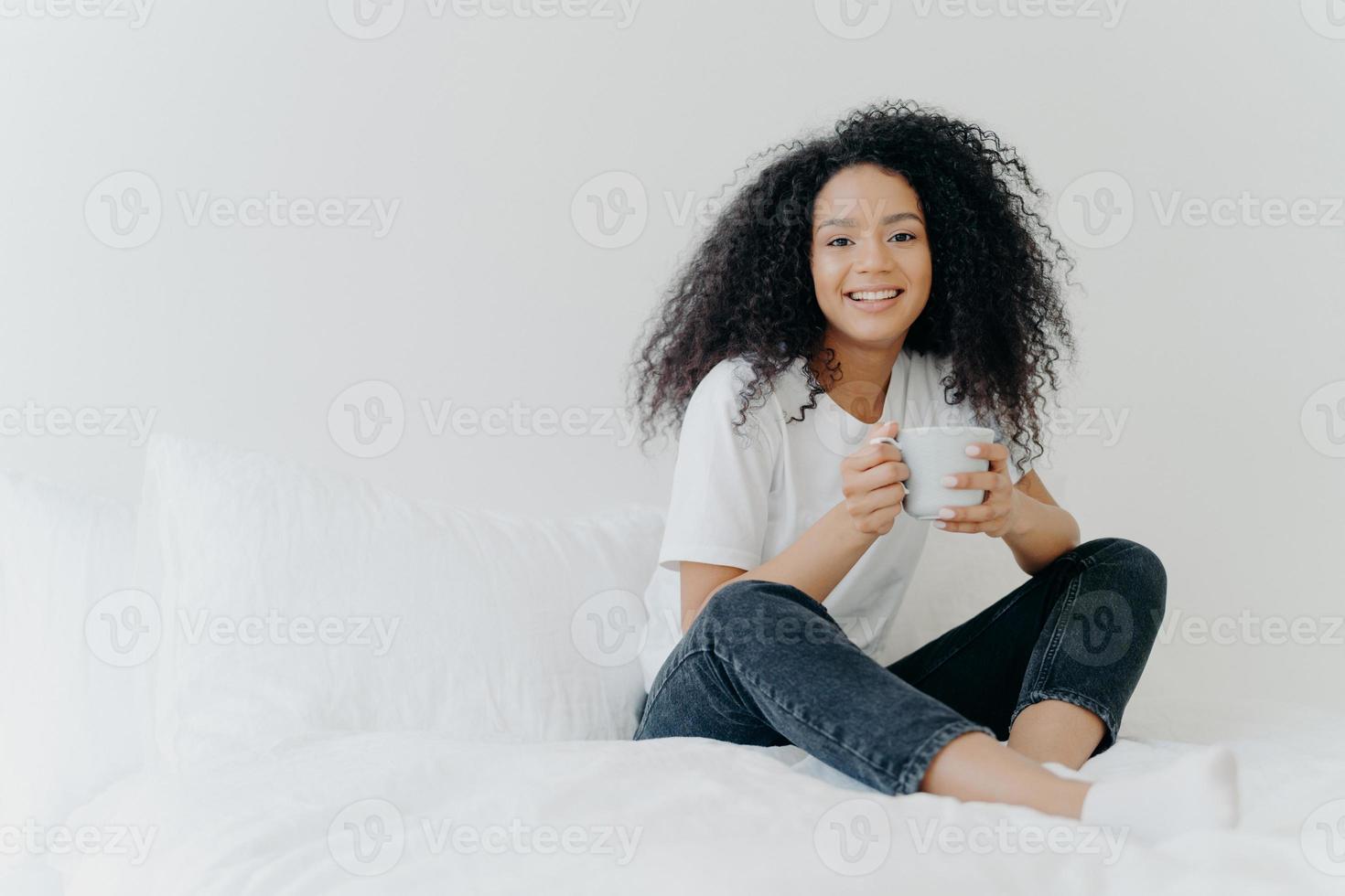 Young pretty African American woman rests in bed at home, drinks hot tea in morning, enjoys domestic atmosphere, tasty drink while relaxes in bedroom. People, leisure, rest and lifestyle concept photo