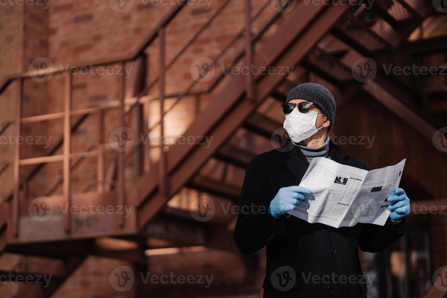 foto horizontal de un hombre pensativo posa con el periódico, usa una máscara protectora y guantes de goma, mira a un lado, piensa en el autoaislamiento, la cuarentena y la pandemia del coronavirus. personas y covid 19