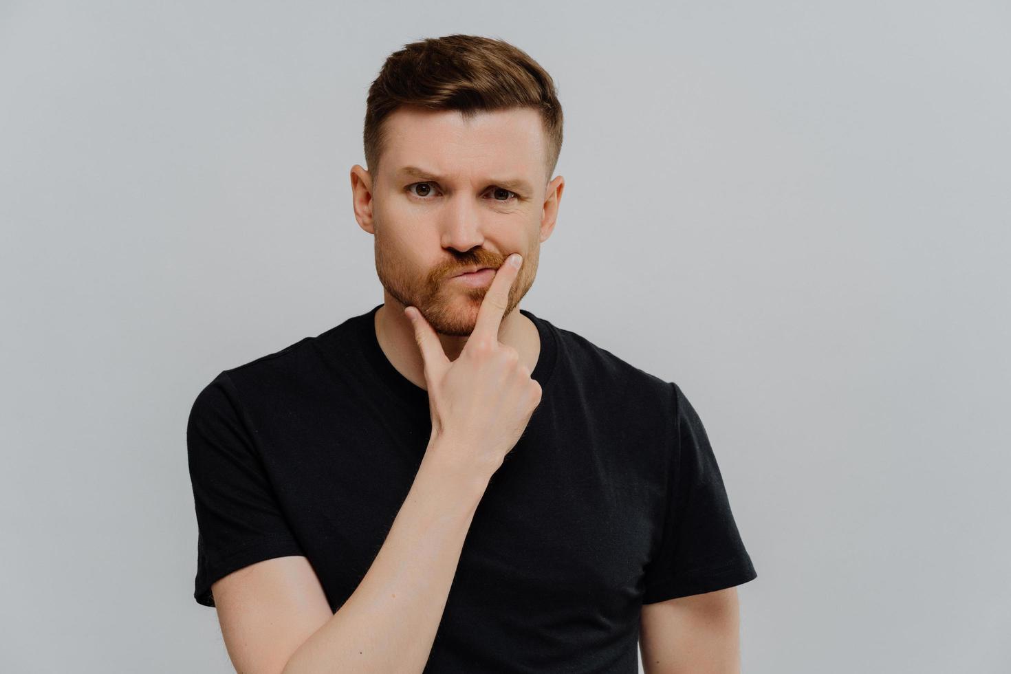 Studio shot of serious displeased man holds chin purses lips looks with thoughtful expression poners on something tries to make up his mind dressed in casual clothes isolated over blue background photo