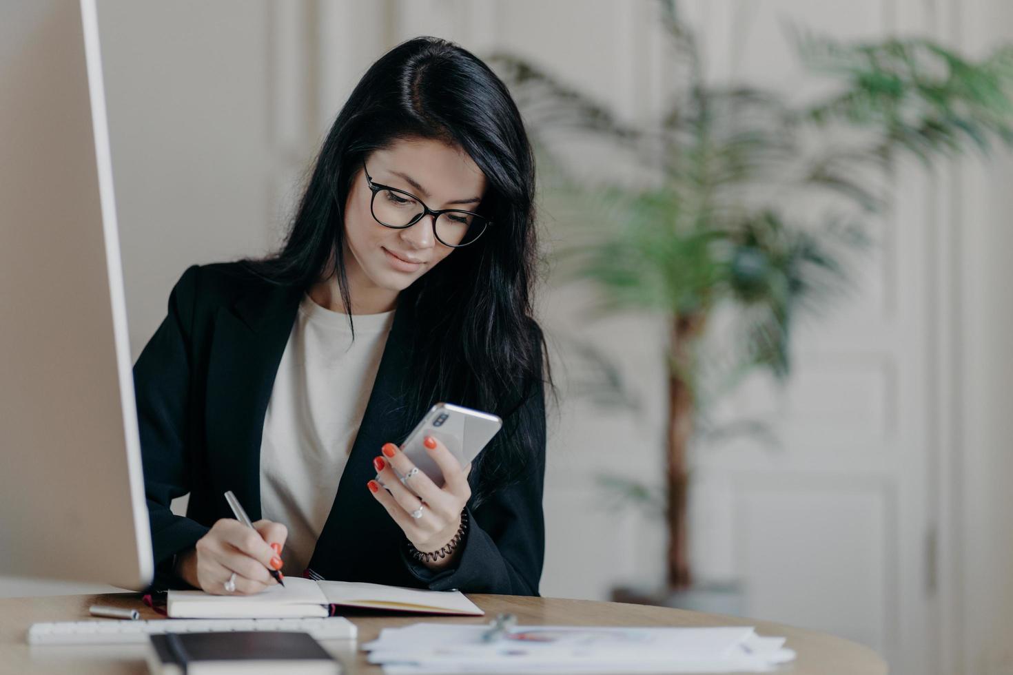 Serious good looking adult woman with dark hair, concentrated into mobile phone, searches necessary information in internet, writes down notes in notepad, sits at desktop, surrounded by papers photo