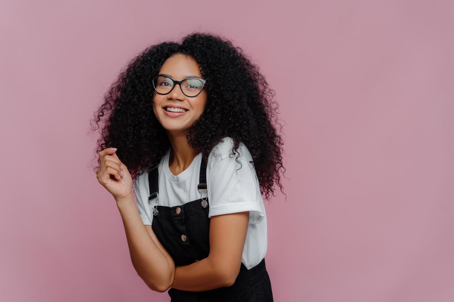 Studio shot of happy dark skinned lady with crisp hair, has dreamy expression, wears optical glasses, stylish clothes, imagines something pleasant, stands against bright violet background, free space photo