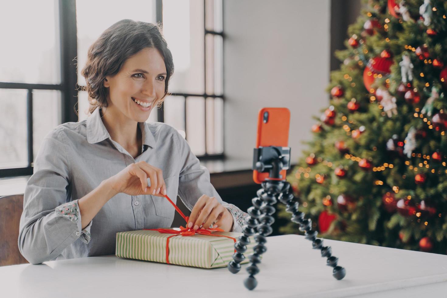 Joyful lady sitting at table with Xmas tree on background in front of phone on tripod photo