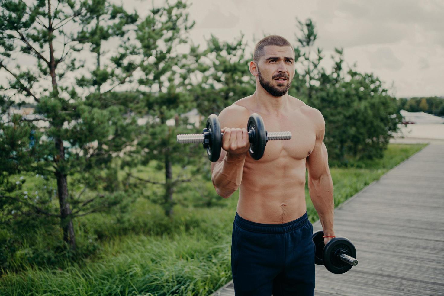 Body care and power lifting concept. Strong muscular athlete bearded man raises dumbbells, lifts heavy barbells, performs shoulder press, poses outside against nature background. Sport exercising photo