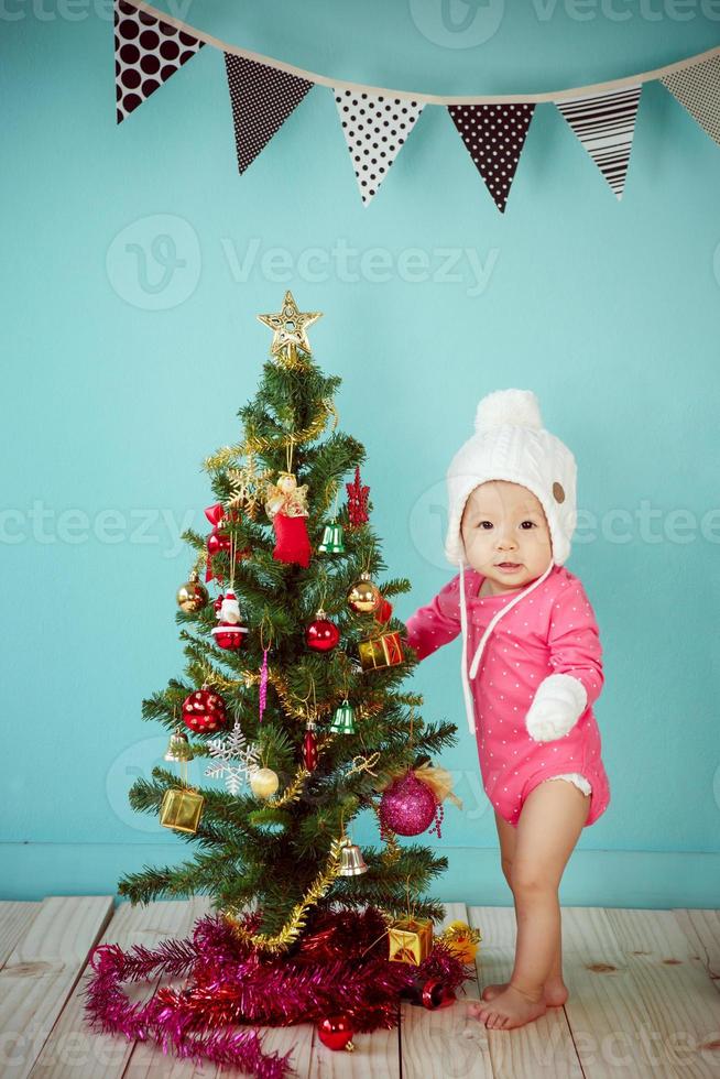 bebé con gorro de punto blanco delante de un fondo azul y decorando un árbol de Navidad con fondo verde foto