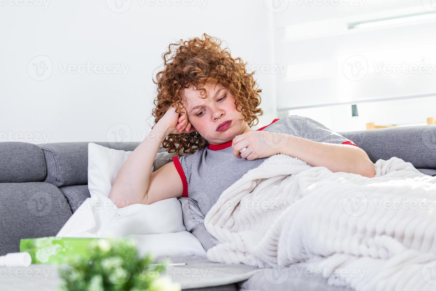 Weak woman, wearing casual clothes is sitting on a sofa and taking a pill. Sick day at home. Young woman has common cold. Sickness, seasonal virus problem concept. photo