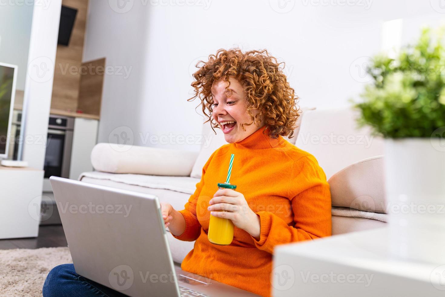 Young ginger girl having video call via laptop with her family while in quarantine. Young student abroad having video call conversation from her laptop at home and drinking an healthy orange juice. photo