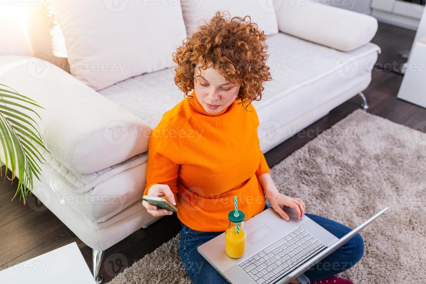 Cute female red hair freelancer reading text messages while sitting on floor with open computer in modern home, young creative woman work on laptop while having orange juice photo
