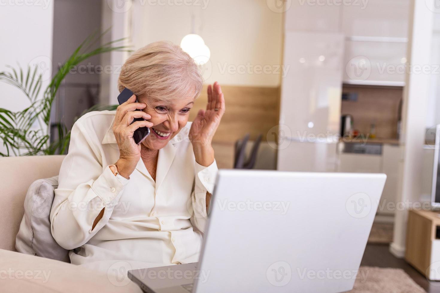 Happy senior Woman with Laptop Computer, Sitting at the Couch While Talking to Someone on Phone. Using from new technology is easier and faster photo