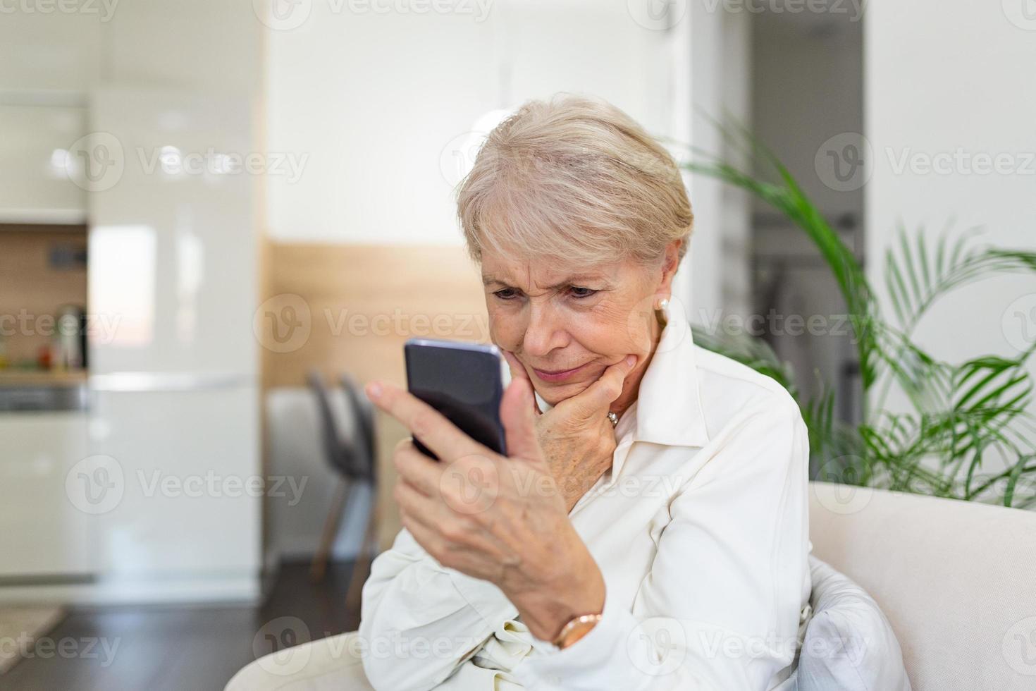 mujer mayor confundida que tiene problemas para usar el teléfono móvil en casa. anciana de pelo blanco sentada en el sofá y tratando de enviar mensajes con un smartphone. foto