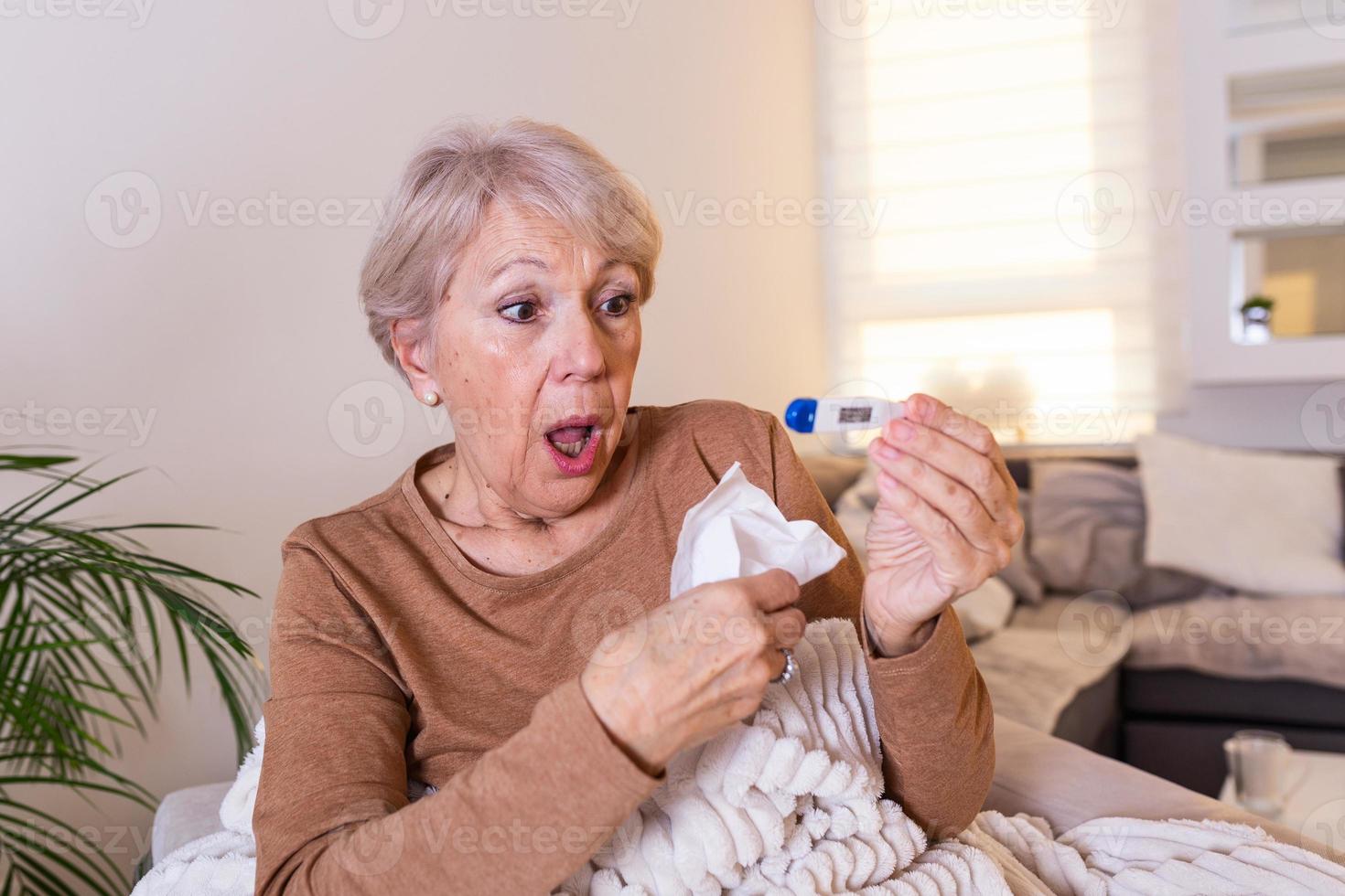 Sickness, seasonal virus problem concept. Senior woman being sick having flu lying on sofa looking at temperature on thermometer. Sick older lady lying in bed with high fever. Cold flu and migraine. photo