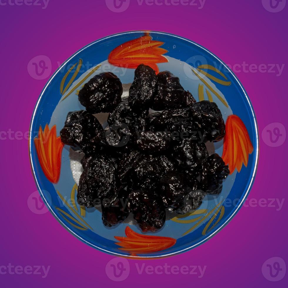 Dried black plums in a glass bowl. Isolated on purple gradient. Top view photo