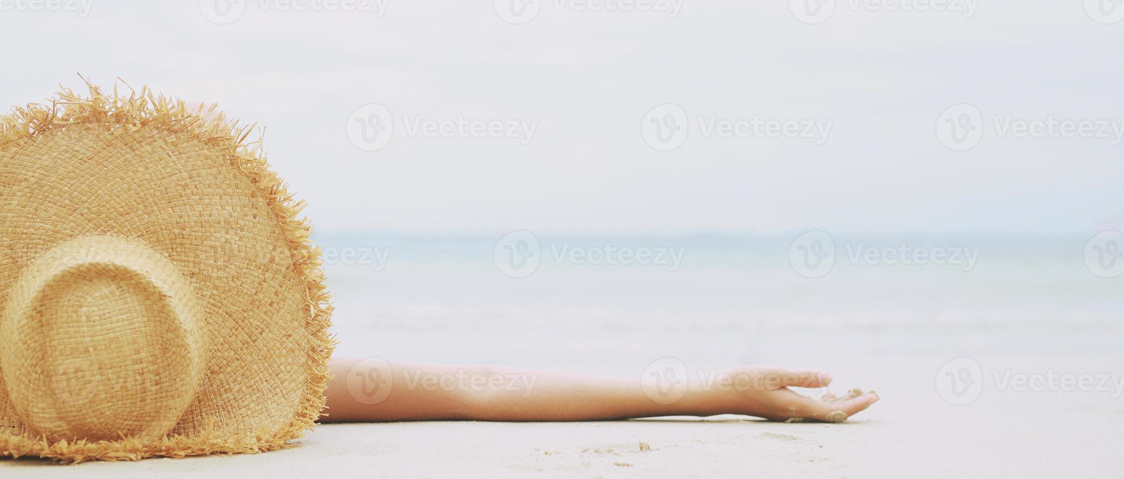 tanning suntanned young woman in bikini wearing straw hats lying on a tropical beach on white sand stretching up slender leg Blue sea in the background. Summer holiday fashion vacation travel concept. photo