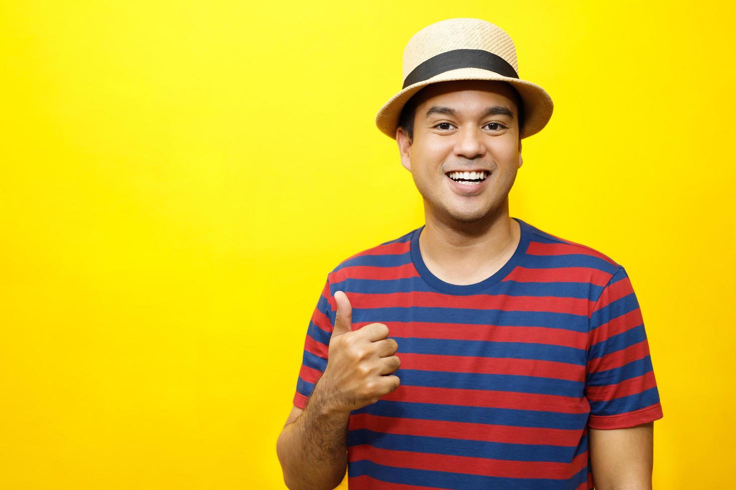 Portrait of cheerful asian man in basic clothing Casual wear red striped T-shirt and wear a hat smiling and show giving thumbs up at camera with showing success. isolated over yellow background. photo