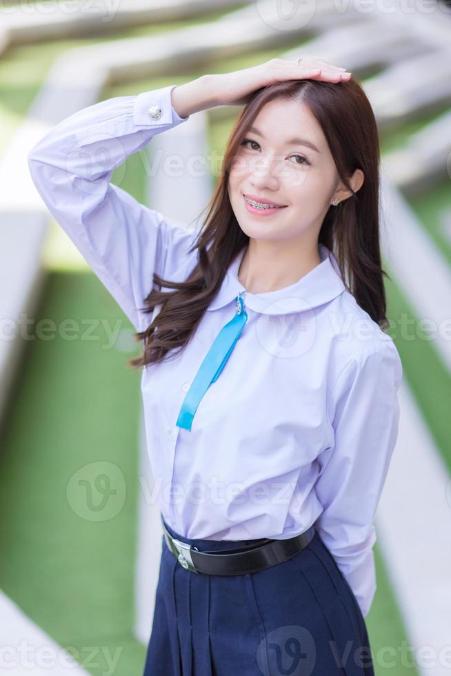 Beautiful high school Asian student girl in the school uniform stands and smiles happily with braces on her teeth while she put her hand on head confidently with the building as a background. photo