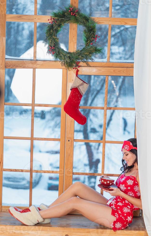 Beautiful young brunette woman wearing red pajamas sitting home by the window photo