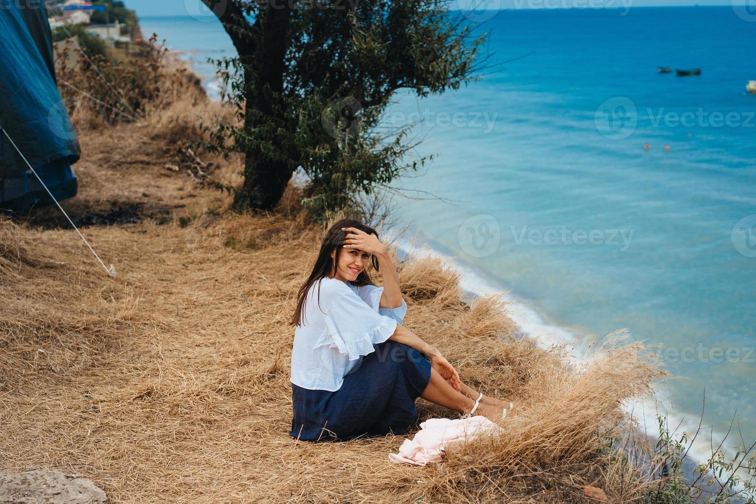 attractive woman in summer skirt and shirt sits on the shore photo