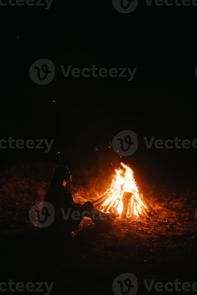 mujer sentada y calentándose cerca de la hoguera en el bosque nocturno. foto