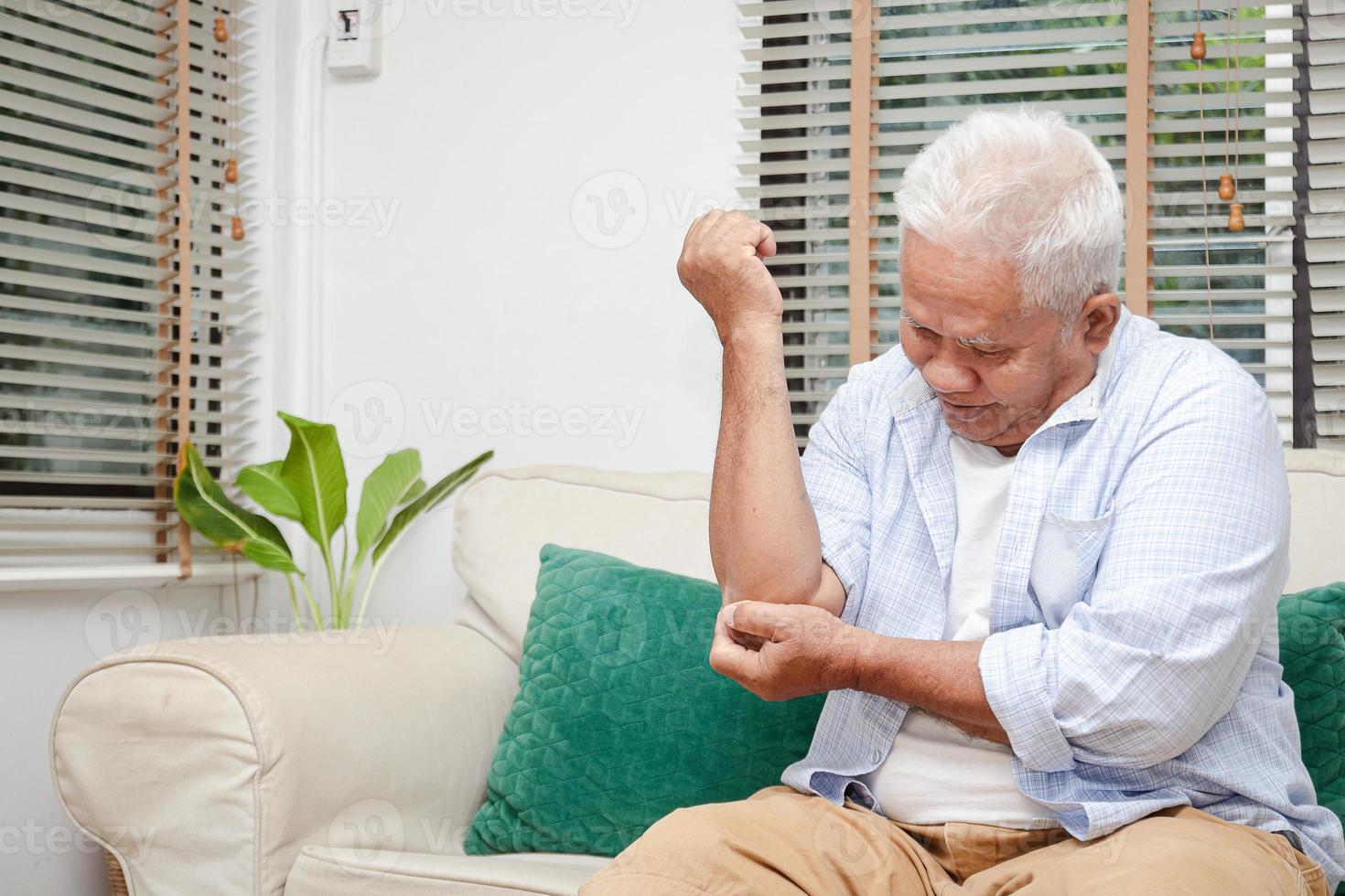 An elderly Asian man sits with arm pain on the sofa in his house. Concept of treatment of osteoarthritis, aging disease. Copy space photo