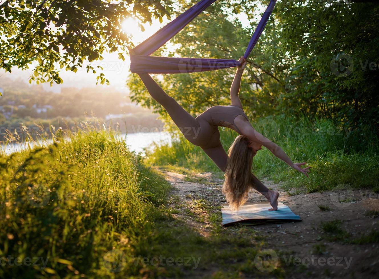 una joven gimnasta se dedica al yoga aéreo en la naturaleza en el parque, usando una combinación de posturas tradicionales de yoga, pilates y danza suave foto