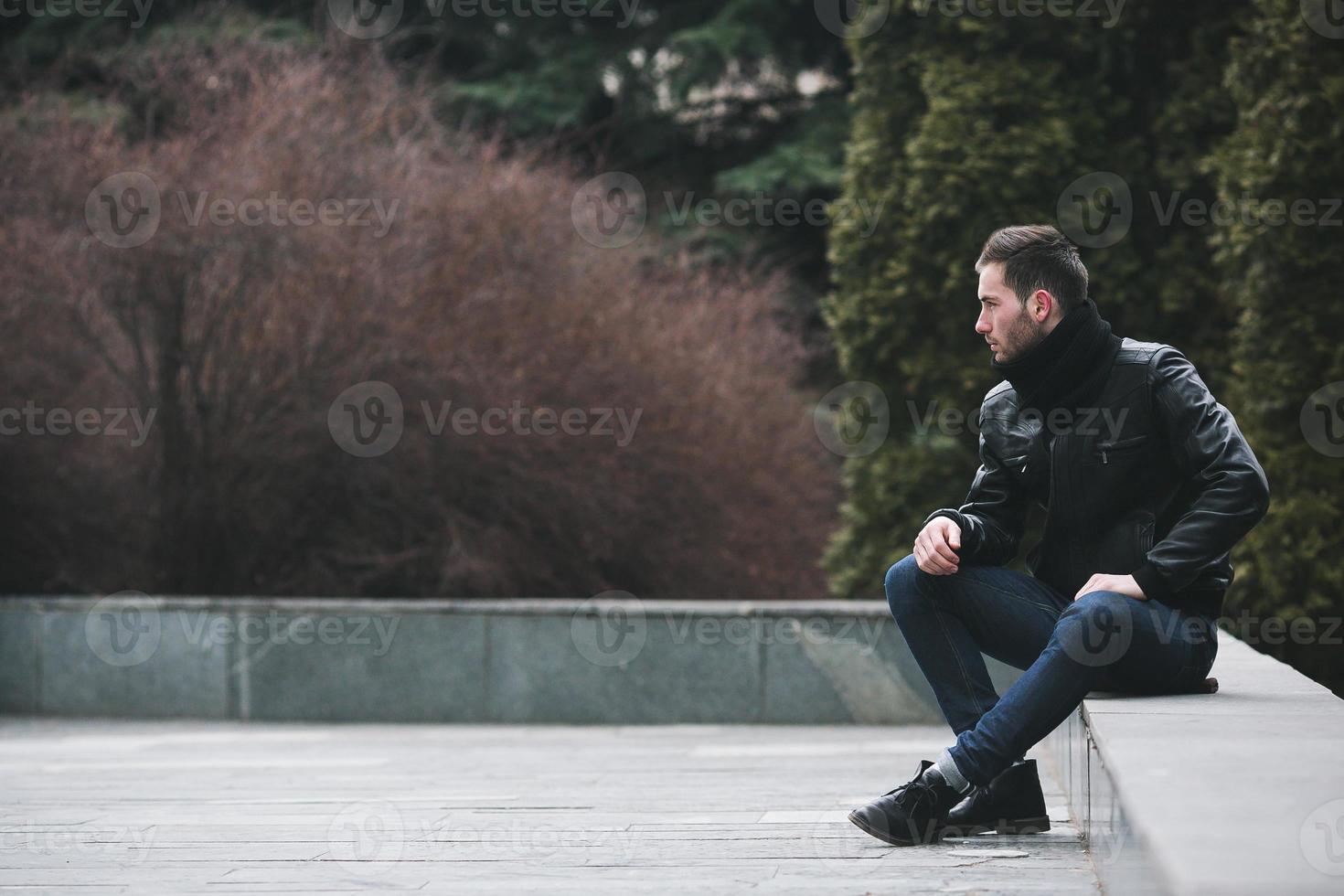 A man dressed in jeans and black jacket seats on a slab photo