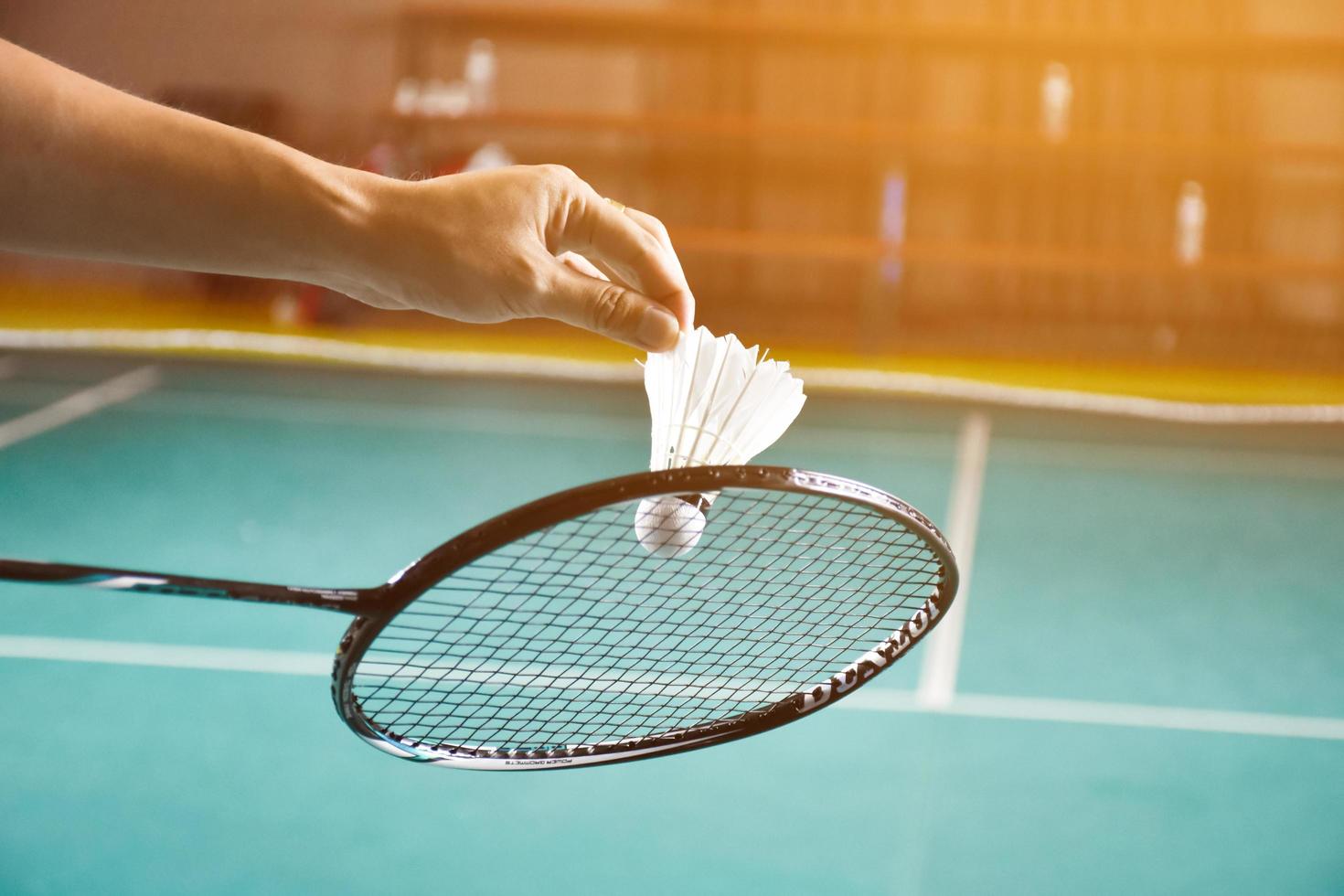 Badminton racket and old white shuttlecock holding in hands of player while serving it over the net ahead, blur badminton court background and selective focus photo