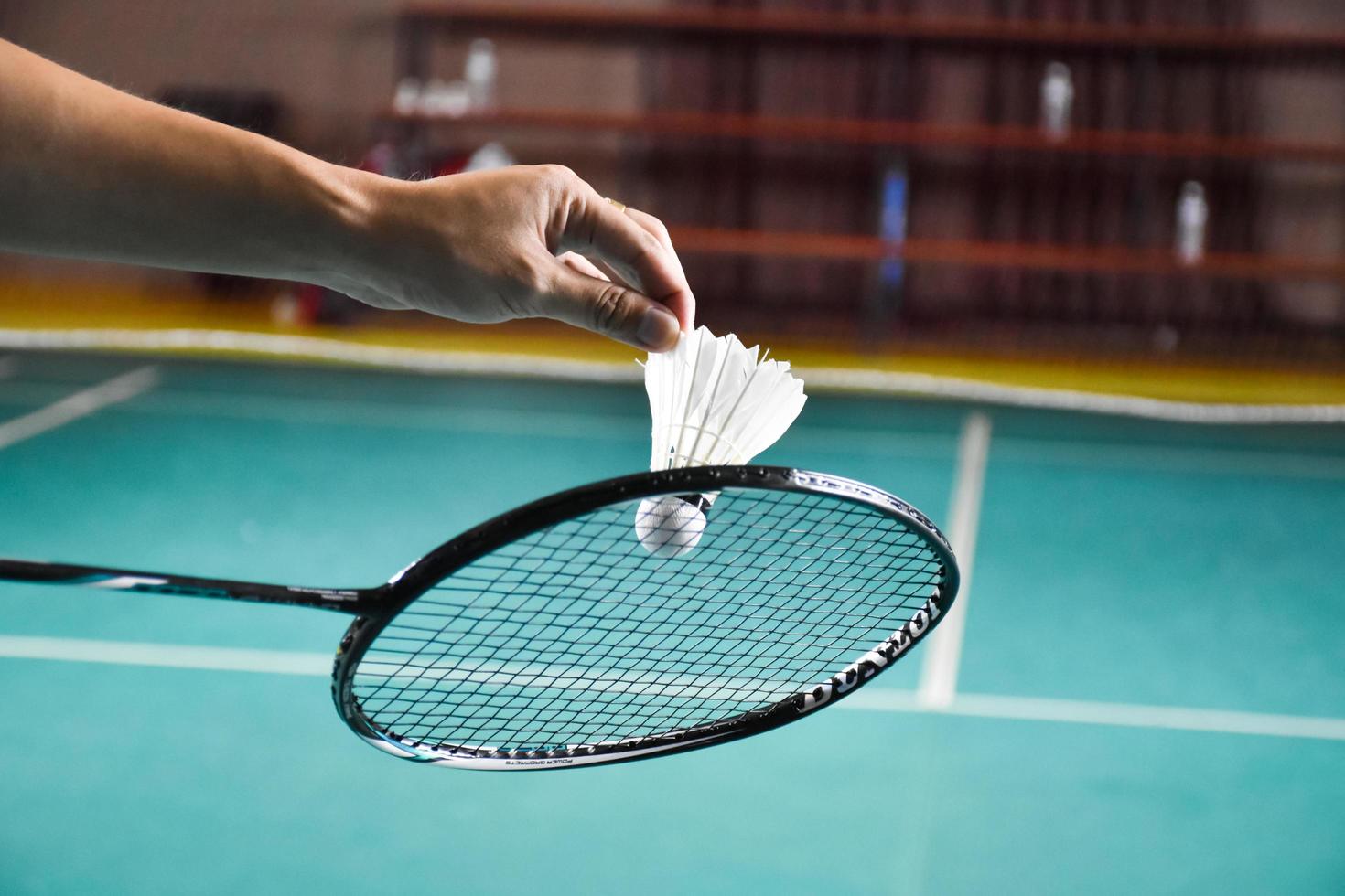 Badminton racket and old white shuttlecock holding in hands of player while serving it over the net ahead, blur badminton court background and selective focus photo