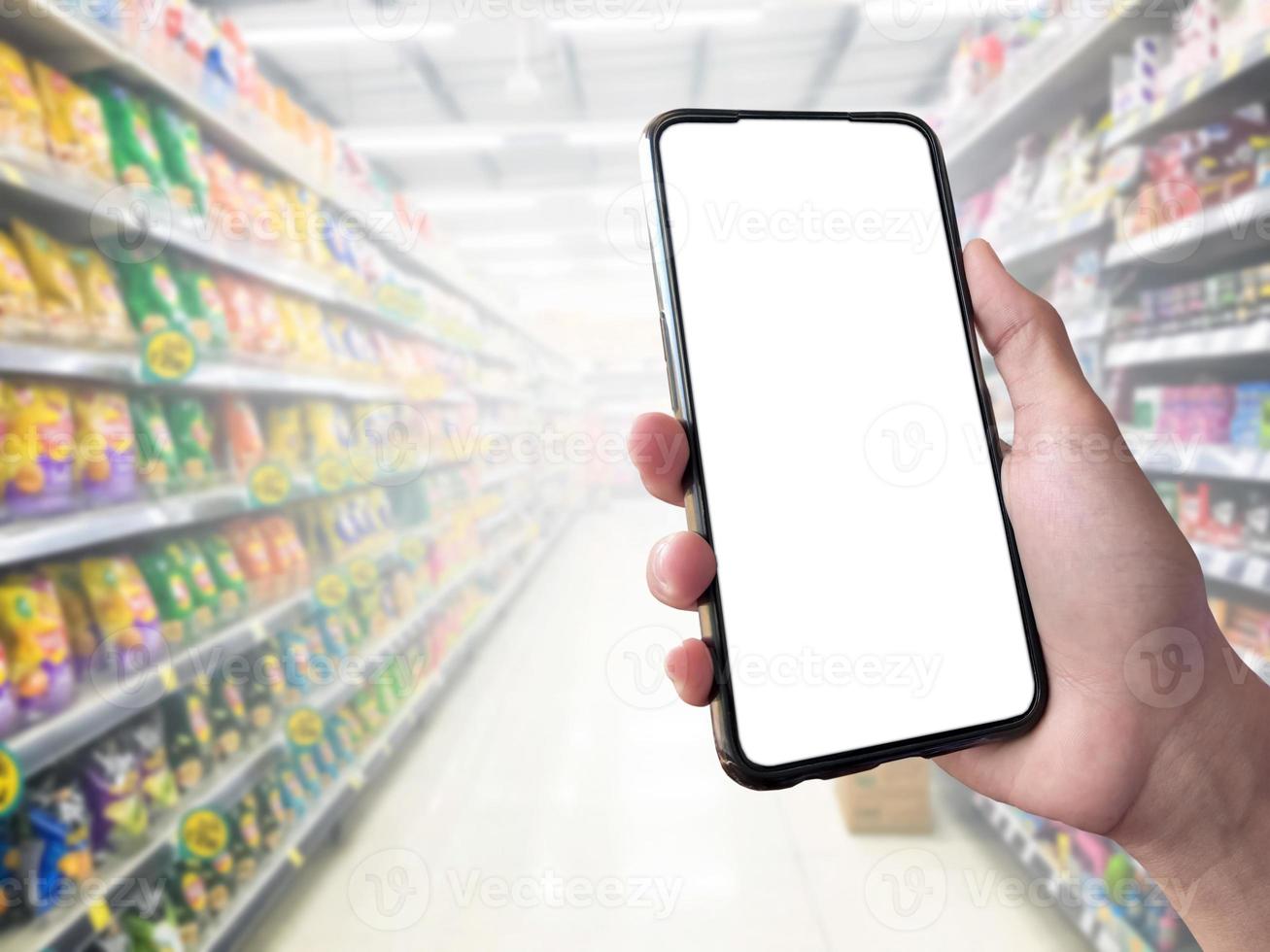 concepto de compra inteligente teléfono móvil en la mano frente a estantes de productos en supermercados y tiendas de comestibles. maqueta de pantalla blanca en blanco para su propia creatividad. foto