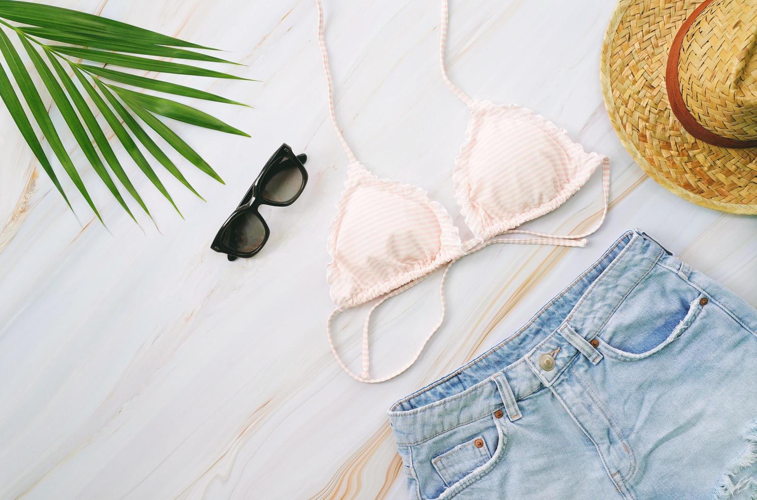 Flat lay of summer items with pastel bikini, sunglasses, jeans, hat and green tropical plant  on marble background, fashion and summer concept photo