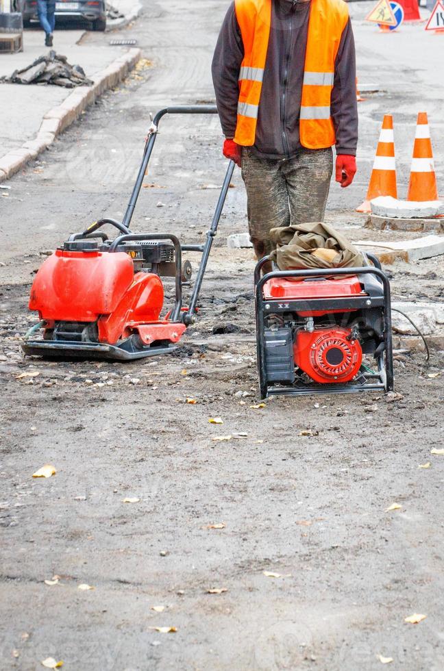 A road worker in an orange vest is repairing the roadway using a gasoline generator and a plate compactor. photo