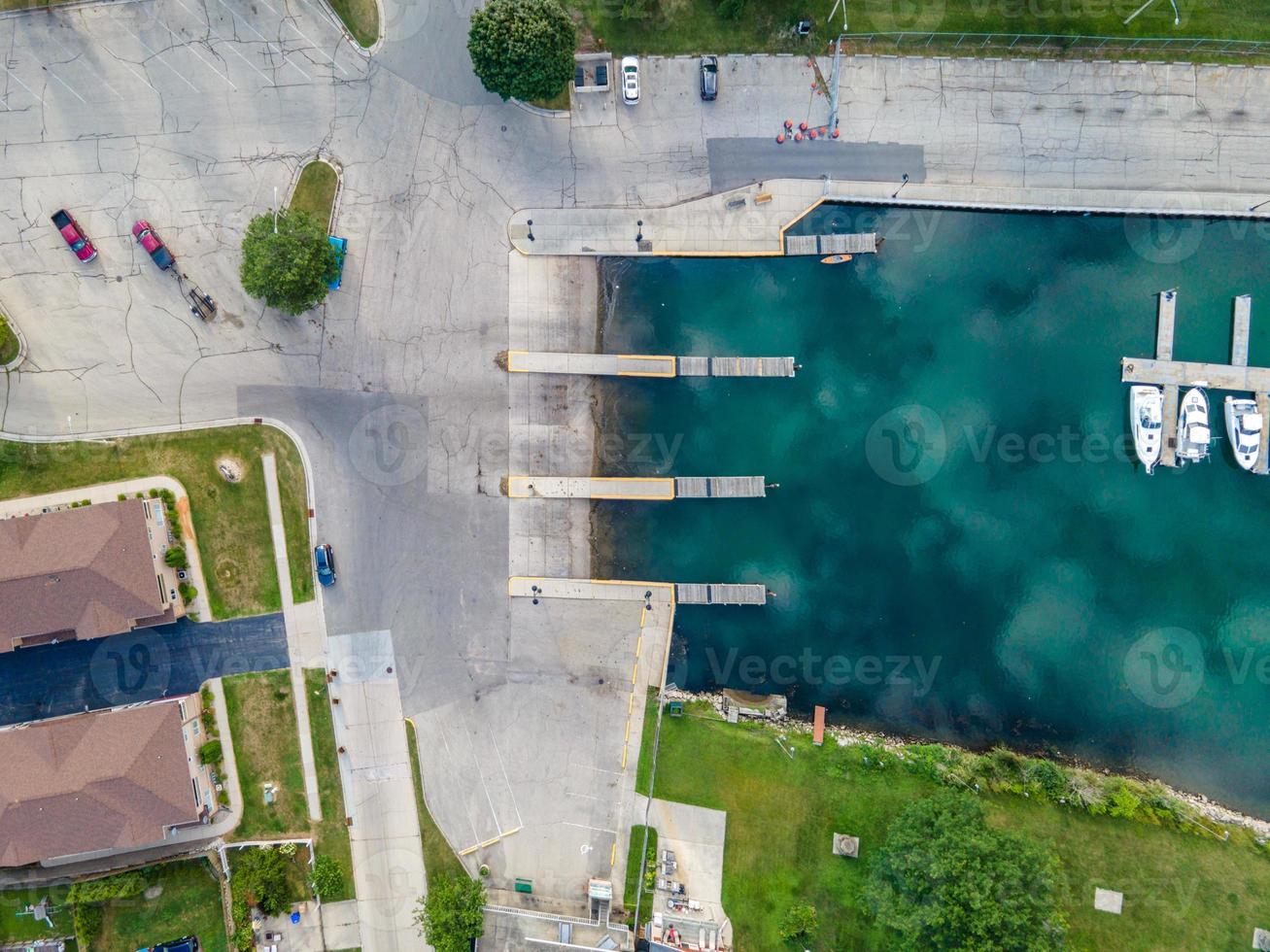 Top down view of boat dock with shadows from the sun. Varied blue patterns in the water. Boat pier with docked boats. Several vehicles in parking lot. Grass along both sides. Residential buildings. photo
