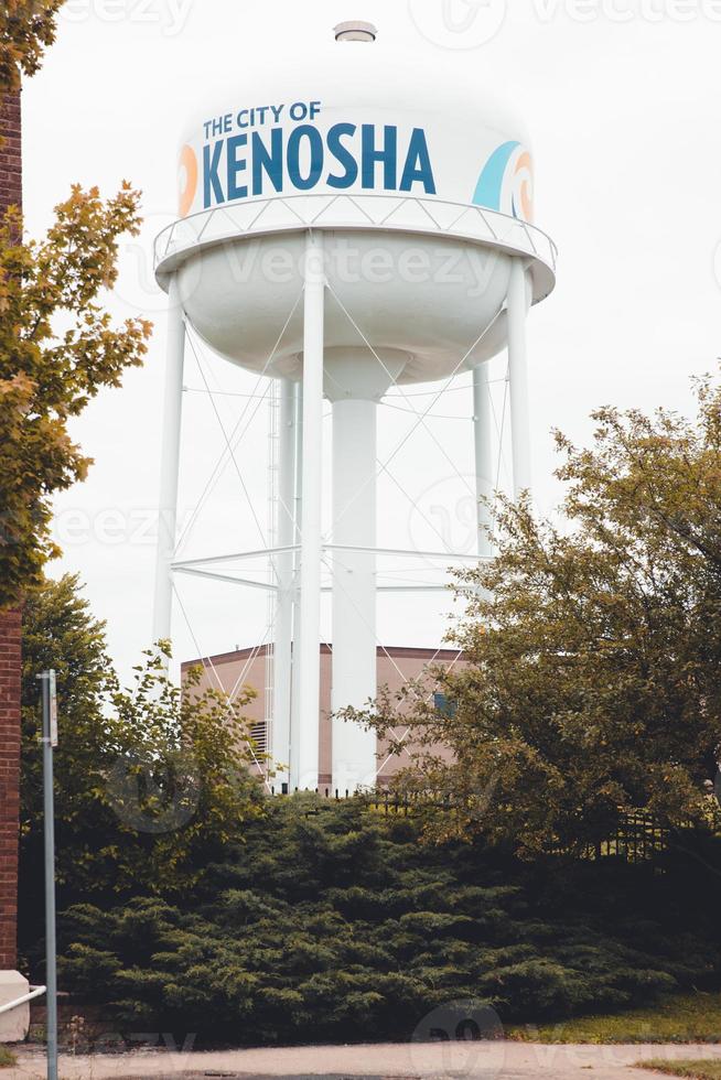 Kenosha, Wisconsin, water tower with  trees around the base. Cloudy sky. Fence hidden by the bushes and trees. Brick building to one side. Blue letters on the tower. Sidewalk in front. photo