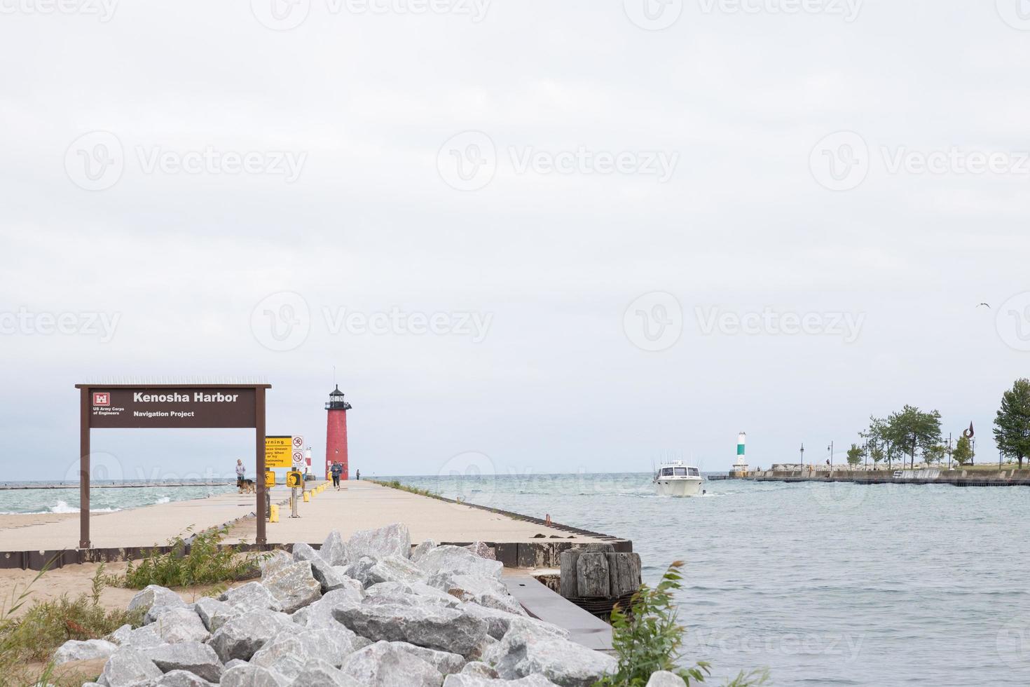 View out Kenosha Harbor on a windy cloudy day. People on the pier.  A boat coming back from the Lake Michigan. Waves crashing into the pier. Boulders along shoreline of pier. photo