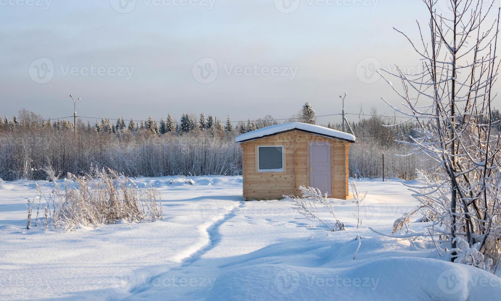 Wooden house at edge of forest. One-storey cottage in winter weather. Single private house near forest. Country house. Trail goes to cottage in middle of snowdrifts. Cottage on a sunny winter day. photo